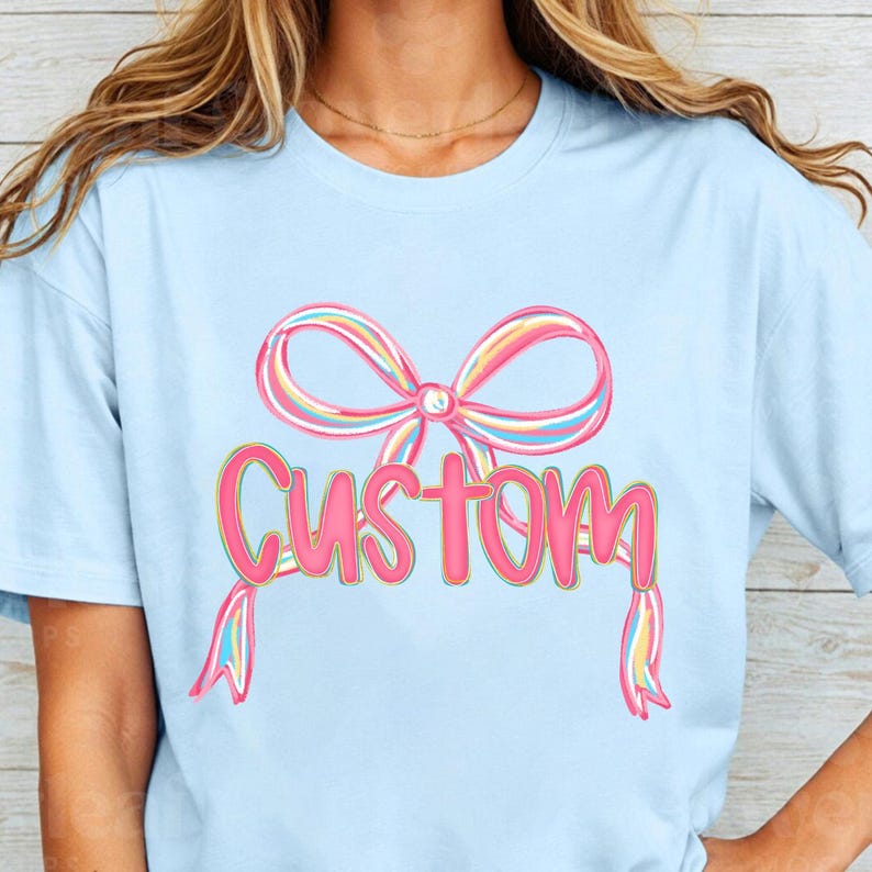 May include: Pale blue t-shirt with a colourful bow and the word "Custom". The bow is striped with pink, yellow, and blue. The word "Custom" is pink with a yellow outline, in a whimsical font.