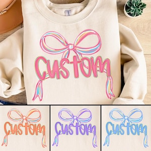 Op de afbeelding: Crèmekleurige sweatshirt met een grote, kleurrijke strik en het woord "Custom" in een speels lettertype. De strik heeft pastelkleurige regenboogstrepen en het woord "Custom" is in een heldere roze tint. Het ontwerp wordt herhaald in drie andere kleuropties.