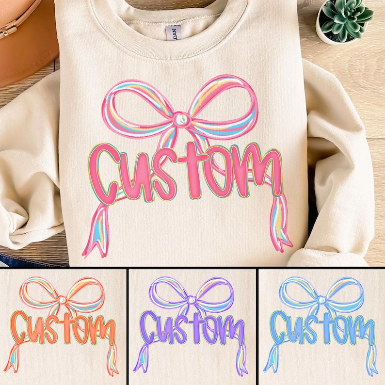 May include: A cream-coloured sweatshirt featuring a large, colourful bow and the word "Custom" in a playful font. The bow has pastel rainbow stripes, and the word "Custom" is in a bright pink hue. The design is repeated in three other colour variations.