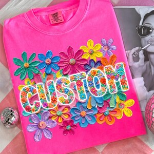 May include: A vibrant pink t-shirt featuring the word "CUSTOM" in a floral design. The word is surrounded by colourful, three-dimensional flowers in shades of blue, pink, yellow, orange, and green. The shirt has a crew neck and short sleeves.