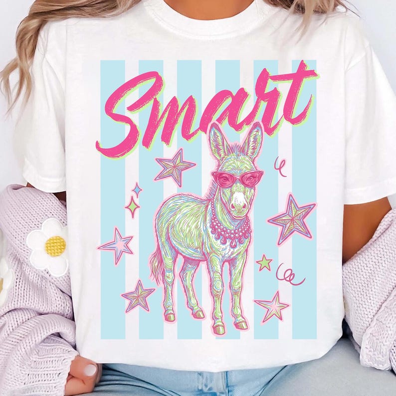 Pode incluir: Camiseta branca com uma ilustra&ccedil;&atilde;o extravagante de um burro usando &oacute;culos de sol rosa e um colar, cercado por estrelas. A palavra "Smart" est&aacute; escrita acima do burro em script rosa, em um fundo de listras azuis e brancas.