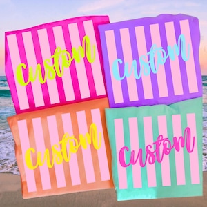 K&ouml;nnte beinhalten: Vier farbenfrohe Strandt&uuml;cher mit vertikalen rosa und wei&szlig;en Streifen. Jedes Handtuch tr&auml;gt das Wort "Custom" in einer anderen Farbe: Neongr&uuml;n, Hellblau, Gelb und Pink. Die Handt&uuml;cher sind an einem Strand angeordnet.
