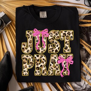 May include: Black t-shirt with the words "JUST PRAY" in leopard print with gold glitter trim and pink bow accents. The shirt is a Comfort Colors brand.