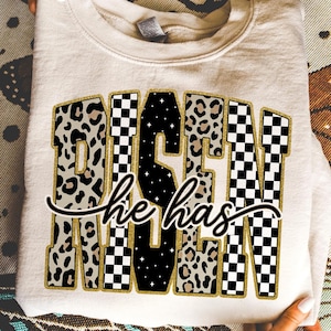 May include: Cream-coloured sweatshirt featuring the text "DESIGN he has" in a mix of leopard print, black and white chequered, and black with white star patterns. The text is outlined in gold glitter.