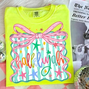 May include: A neon yellow Comfort Colors t-shirt with a colorful graphic. The graphic features a pink bow, teal and white stripes, colorful stars, and the word "hallelujah" in a rainbow of colors. The shirt is a size L-G.