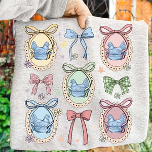 May include: A light gray sweatshirt featuring a repeating pattern of blue hen illustrations inside oval frames with decorative bows. The bows are in various colors, including yellow, pink, and green, with floral accents.