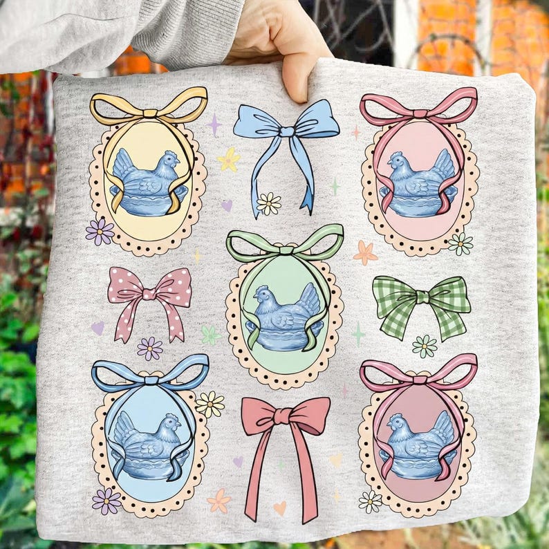 May include: A light gray sweatshirt featuring a repeating pattern of blue hen illustrations inside oval frames with decorative bows. The bows are in various colors, including yellow, pink, and green, with floral accents.