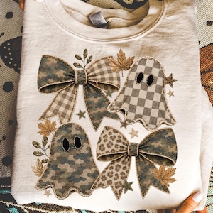 May include: A cream-colored crewneck sweatshirt featuring a graphic print of two ghosts and two large bows. The design incorporates camouflage, checkered, and leopard patterns in muted earth tones, accented with small stars and maple leaves.