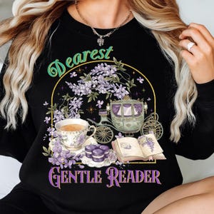 May include: Black sweatshirt with a whimsical design featuring a teacup, carriage, and open book. The text "Dearest Gentle Reader" is displayed in a decorative font, with floral and purple accents. The design evokes a vintage, romantic theme.
