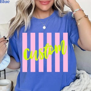 K&ouml;nnte beinhalten: Blaues T-Shirt mit vertikalem rosa Streifendesign und dem Wort "Custom" in neongr&uuml;ner Schrift. Das Shirt ist einfarbig blau und hat einen Rundhalsausschnitt.
