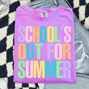 May include: A lilac Comfort Colors t-shirt with the text "SCHOOL'S OUT FOR SUMMER" in colorful block letters. The shirt is folded and rests on a black and white patterned surface, with a pair of blue jeans in the background.