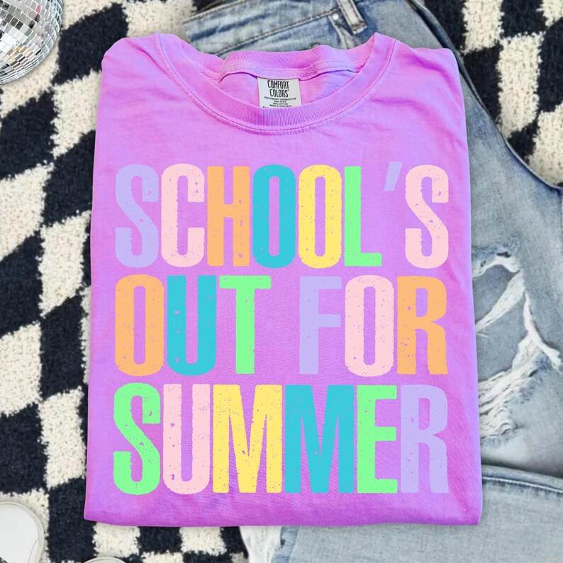 May include: A lilac Comfort Colors t-shirt with the text "SCHOOL'S OUT FOR SUMMER" in colorful block letters. The shirt is folded and rests on a black and white patterned surface, with a pair of blue jeans in the background.