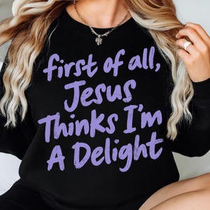 May include: Black sweatshirt with the text "first of all, Jesus Thinks I'm A Delight" in purple script. The sweatshirt is a crew neck style. A gold necklace with a small pendant is visible.