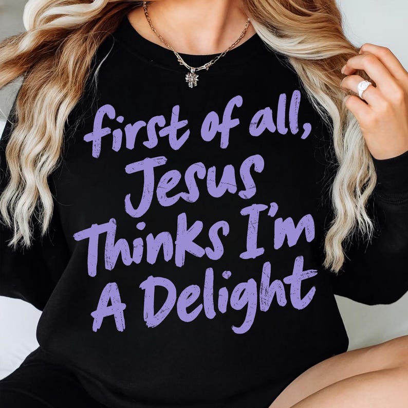 May include: Black sweatshirt with the text "first of all, Jesus Thinks I'm A Delight" in purple script. The sweatshirt is a crew neck style. A gold necklace with a small pendant is visible.