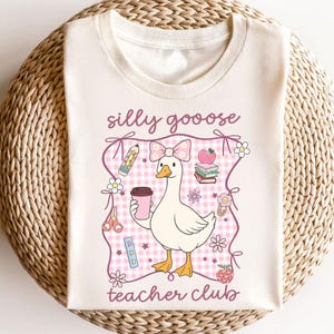 May include: Cream-colored t-shirt featuring a cartoon goose with a pink bow, holding a coffee cup. The design includes the text "silly goooose teacher club" and school-themed graphics like pencils, books, and flowers.