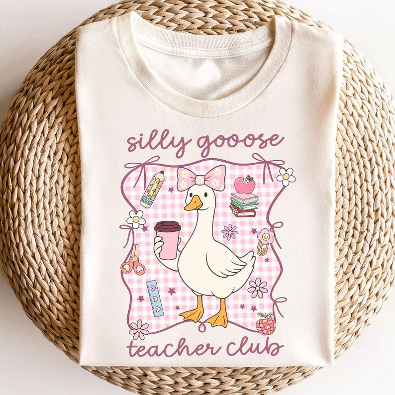 May include: Cream-colored t-shirt featuring a cartoon goose with a pink bow, holding a coffee cup. The design includes the text "silly goooose teacher club" and school-themed graphics like pencils, books, and flowers.