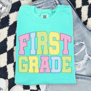 May include: A bright turquoise crewneck t-shirt featuring the text FIRST GRADE in large, block-style varsity letters. Each letter has a vertical striped pattern in alternating colors of pink, blue, yellow, and green across two rows.