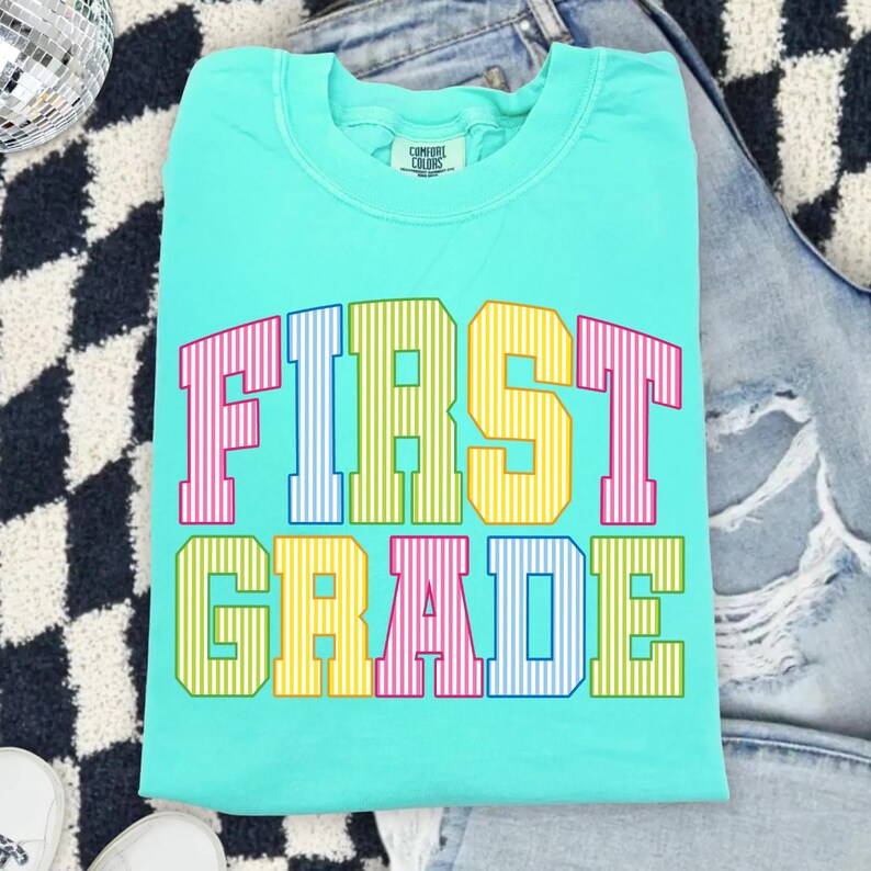 May include: A bright turquoise crewneck t-shirt featuring the text FIRST GRADE in large, block-style varsity letters. Each letter has a vertical striped pattern in alternating colors of pink, blue, yellow, and green across two rows.