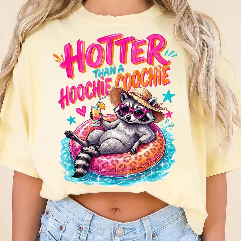 K&ouml;nnte beinhalten: Hellgelbes T-Shirt mit dem Schriftzug "Hotter Than a Hoochie Coochie" in Pink und Orange. Die Grafik zeigt einen Waschb&auml;ren mit Sonnenbrille und Strohhut, der sich auf einem rosa Leoparden-Print-Schwimmring entspannt und einen Cocktail h&auml;lt.