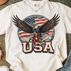 May include: White t-shirt featuring a graphic of an eagle with outstretched wings, wearing sunglasses and a medal. The eagle is set against a backdrop of the American flag and mountains, with the word "USA" in bold letters below.