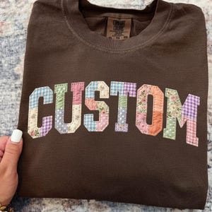 Puede incluir: Una camiseta marrón con la palabra "CUSTOM" aplicada en la parte delantera. Las letras están hechas de tela de patchwork en varios colores y patrones, incluyendo flores, cuadros y lunares. La camiseta tiene cuello redondo y una etiqueta que dice "COMFORT COLORS".