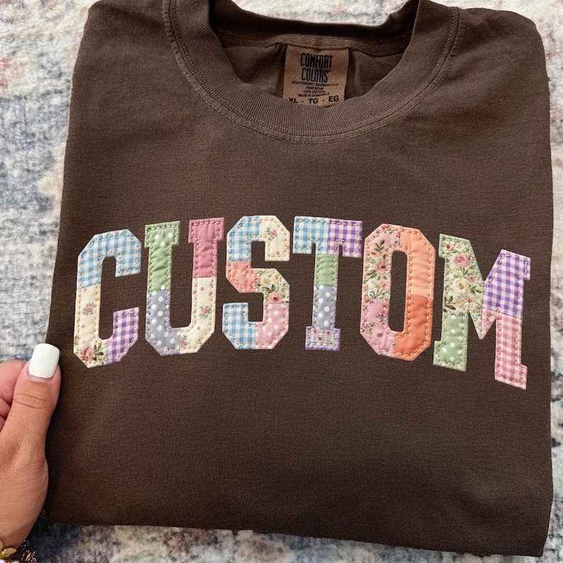 May include: A brown t-shirt with the word "CUSTOM" appliqued across the front. The letters are made of patchwork fabric in various colors and patterns, including floral, gingham, and polka dots. The shirt has a crew neck and a tag that says "COMFORT COLORS".