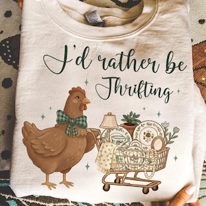 May include: Cream-colored sweatshirt with the text "I'd rather be Thrifting" in dark green script. A cartoon chicken wearing a green plaid bow tie pushes a shopping cart filled with vintage items, including a lamp and dishes.