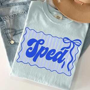 May include: A light blue t-shirt with the word "Spead" in a blue, retro font. The word is inside a blue scalloped border with vertical stripes and a bow. The shirt is folded on top of a pair of blue jeans.