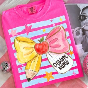 May include: A vibrant pink t-shirt with a watercolour design of a bow, pencil, and apple. The bow is pink and yellow, with a red apple in the centre. The shirt has the words "Custom Name" on a notepad.