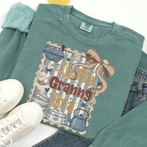 以下が含まれることがあります： 前面に「In My Granny Era」というフレーズがプリントされた、ティール色のスウェットシャツ。イラストには、ガチョウ、ティーカップ、ティーポット、リボンが含まれています。白いスニーカーとデニムジーンズが見えます。