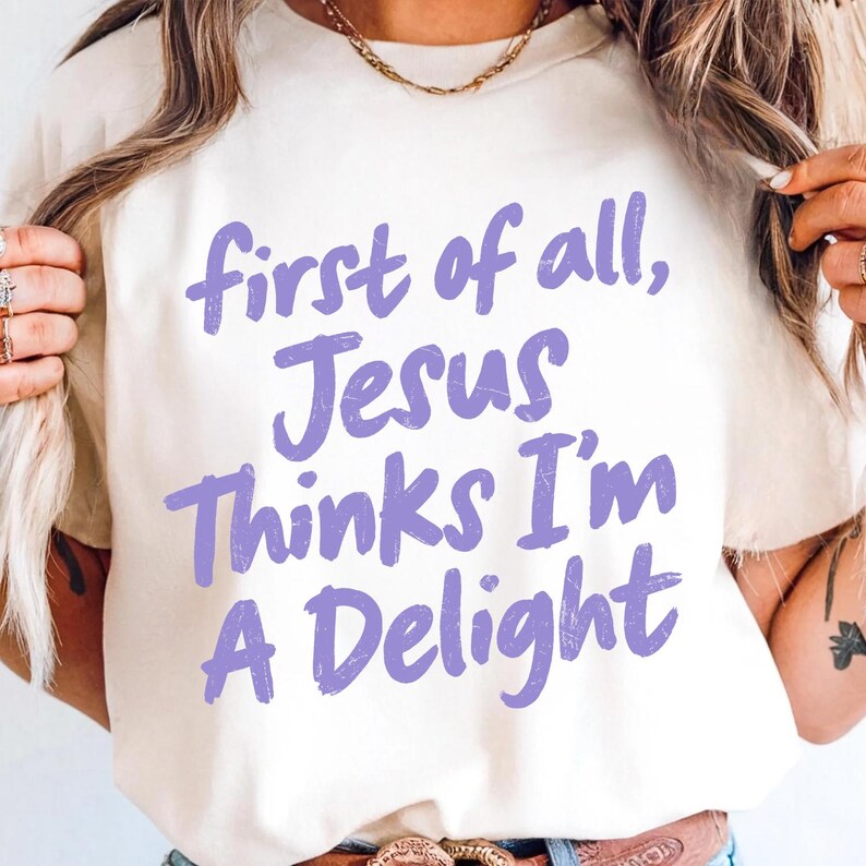 May include: Cream-colored t-shirt with the phrase "first of all, Jesus thinks I'm a delight" in lavender script. The shirt is a casual style, suitable for everyday wear. The text is centered on the front of the shirt.
