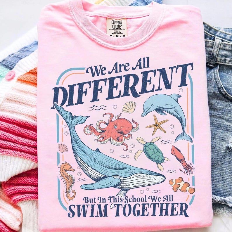 May include: Pink t-shirt with the text "We Are All DIFFERENT But In This School We All SWIM TOGETHER." The graphic features a whale, octopus, dolphin, starfish, seahorse, turtle, squid, and clownfish.