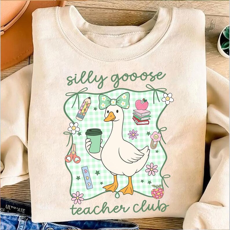 May include: Cream-colored sweatshirt with a whimsical graphic of a goose holding a coffee cup, surrounded by school-themed illustrations. The text "silly goooose teacher club" is displayed in a playful font. The design includes a green gingham background and floral accents.