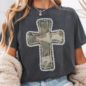 May include: Dark gray t-shirt featuring a large cross design. The cross is made of camouflage fabric, outlined with faux pearls. The shirt has a relaxed fit and short sleeves, suitable for casual wear.