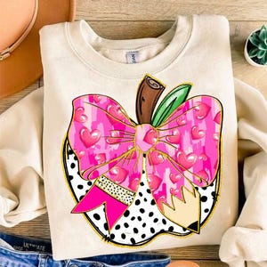 Pode incluir: Uma camisa de cor creme com um desenho de maçã rosa. A maçã tem um laço rosa com corações e um padrão de bolinhas pretas e brancas. A maçã também tem um caule marrom e folhas verdes.