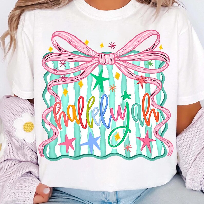 May include: White t-shirt featuring a colorful graphic design. The design includes a pink bow, teal stripes, and the word "Hallelujah" in various colors, along with stars and diamond shapes.