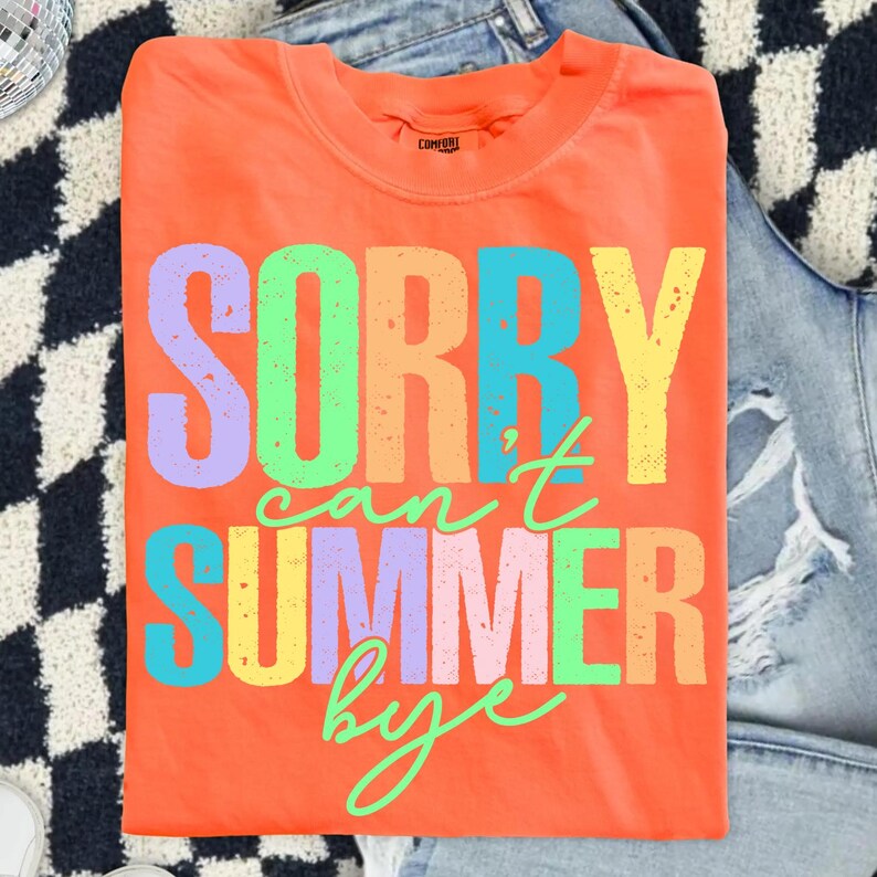 May include: An orange t-shirt with the text "SORRY can't SUMMER bye" in colorful block letters. The shirt is folded and rests on a pair of blue jeans. The text is in shades of purple, green, blue, yellow, orange, and pink.