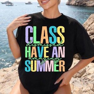 May include: Black t-shirt with the words "CLASS dismissed HAVE AN amazing SUMMER" in colorful, block letters. The shirt has a crew neck and short sleeves. The background shows a seascape with boats.