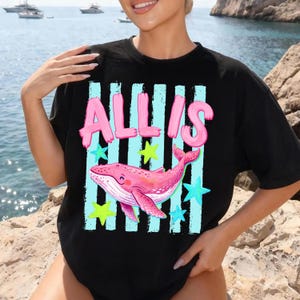 以下が含まれることがあります： ピンクの泡文字で「ALLIS」という名前がプリントされた黒いTシャツ。ピンクのクジラのイラストの上にあります。クジラは青と黄色の星と垂直の青いストライプに囲まれています。