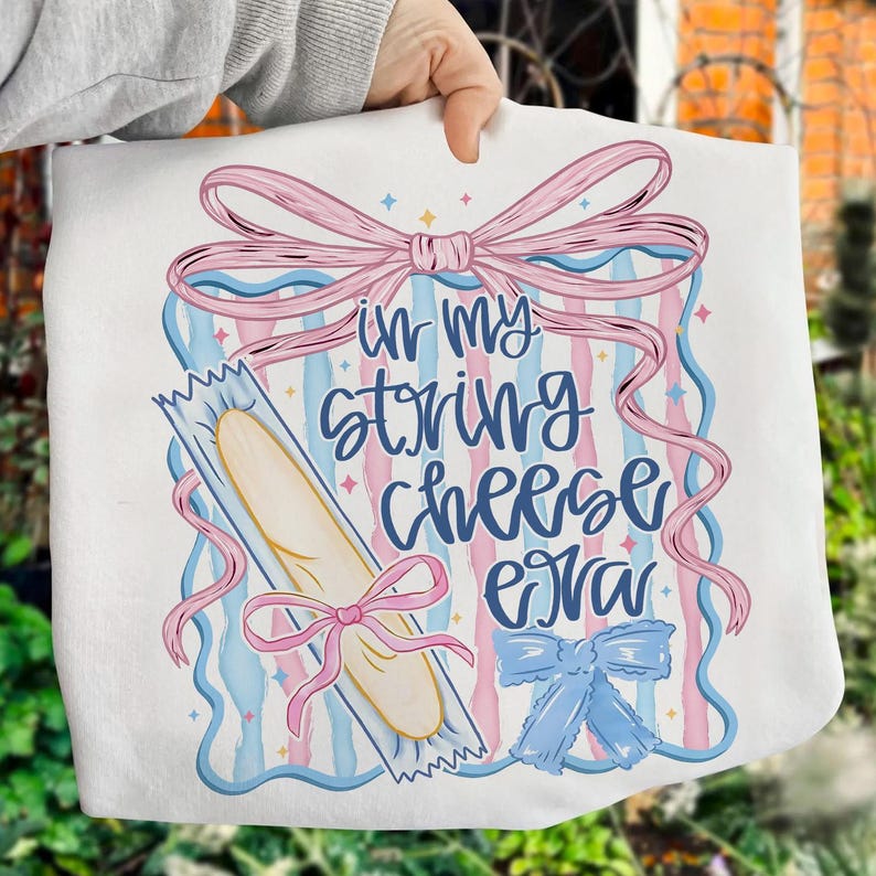 May include: White tote bag with a whimsical design featuring a string cheese illustration and the phrase "in my string cheese era" in blue script. The design includes pink and blue ribbons and stars.
