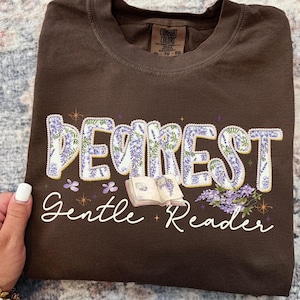 May include: A brown t-shirt with the words "DEAREST Gentle Reader" in a floral design. The text is in white script with purple and yellow floral accents. An open book illustration is also featured.