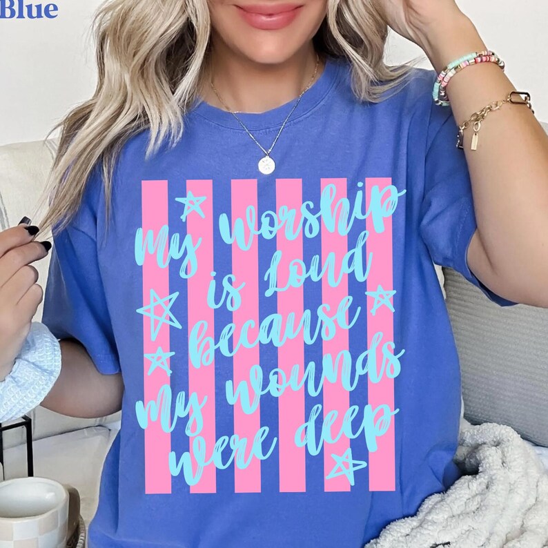 May include: Blue t-shirt with the text "My worship is loud because my wounds were deep" in light blue script and stars, set against pink vertical stripes. The shirt has a crew neck and short sleeves.