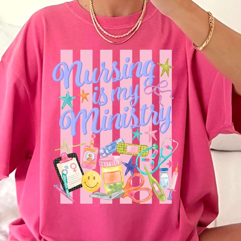 May include: Pink t-shirt with the text "Nursing is my Ministry" in blue script. The design includes medical-themed illustrations such as a stethoscope, pills, and a syringe. The shirt has a striped background.