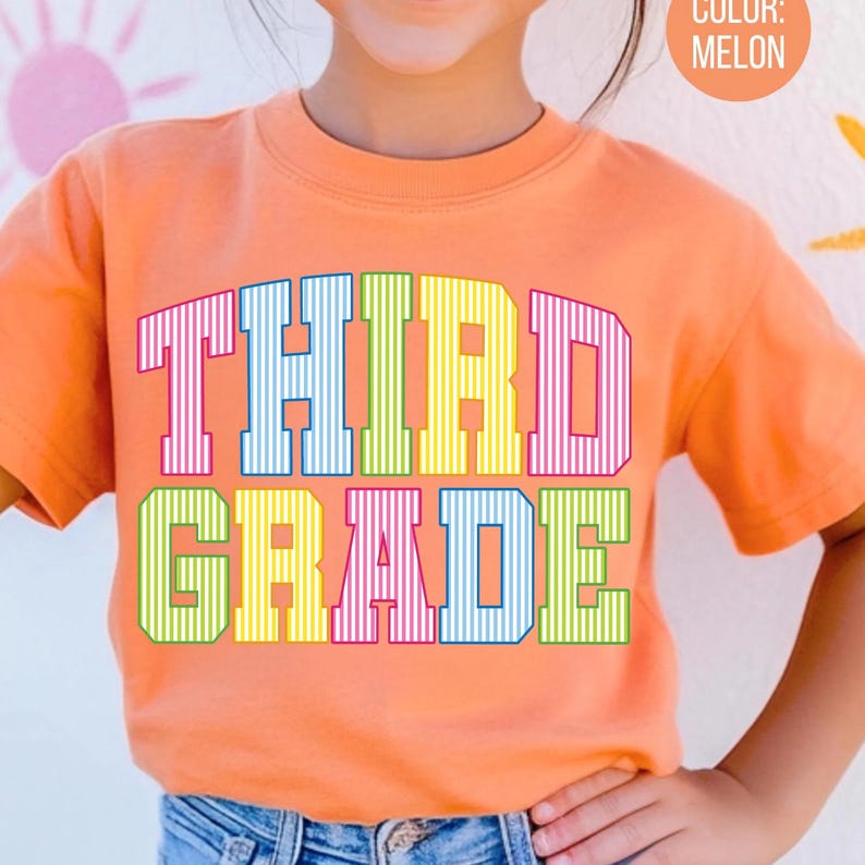 May include: A melon-colored t-shirt featuring the text THIRD GRADE printed in a bold, block-letter font. Each letter is filled with a vertical pinstripe pattern in alternating colors including pink, blue, yellow, and green.