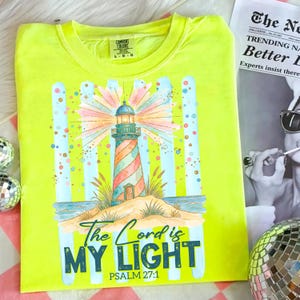 Pu&ograve; includere: Una maglietta Comfort Colors giallo neon con un design a faro colorato. Il faro &egrave; a strisce rosse, arancioni e blu, con una luce brillante che emana dalla cima. Il testo "The Lord is My Light" e il Salmo 27:1 sono stampati sotto.