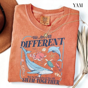 May include: Orange Comfort Colors t-shirt with a marine life graphic and the text "We Are All DIFFERENT But In This School We All SWIM TOGETHER." A gold necklace with a pendant is draped on the shirt.