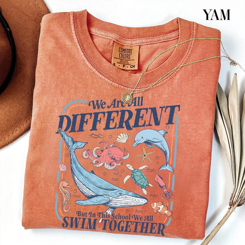 May include: Orange Comfort Colors t-shirt with a marine life graphic and the text "We Are All DIFFERENT But In This School We All SWIM TOGETHER." A gold necklace with a pendant is draped on the shirt.