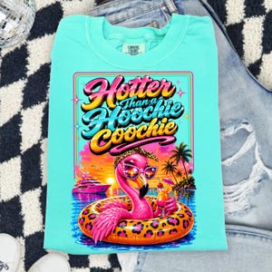 May include: A turquoise Comfort Colors t-shirt featuring a vibrant graphic of a flamingo in sunglasses and a leopard print headband, floating in a pool with a cocktail. The text reads "Hotter Than a Hoochie Coochie."