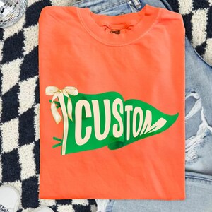 May include: Orange t-shirt with a green pennant design that reads "CUSTOM" in white lettering. A cream-colored bow and ribbon are attached to the pennant. The shirt is folded and placed on a denim surface.