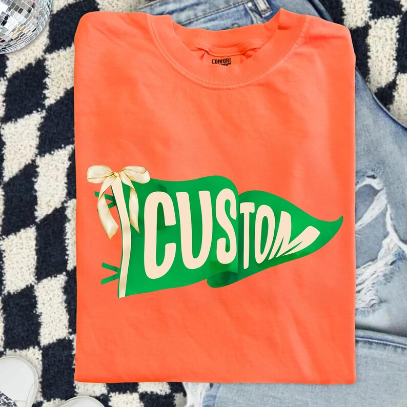 May include: Orange t-shirt with a green pennant design that reads "CUSTOM" in white lettering. A cream-colored bow and ribbon are attached to the pennant. The shirt is folded and placed on a denim surface.